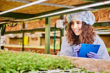 Female agricultural scientist studying plant growth and taking notes in greenhouse. Cheerful woman gardener looking at seedlings and smiling while holding clipboard.