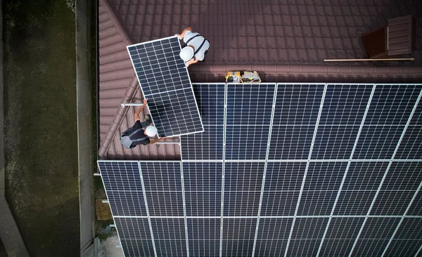 Men technicians mounting photovoltaic solar moduls on roof of house. Workmen in helmets installing solar panel system outdoors. Concept of alternative and renewable energy. Aerial view.