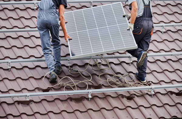 Men installers carrying photovoltaic solar moduls on roof of house. Cropped view of electricians installing solar panel system outdoors. Concept of alternative and renewable energy.