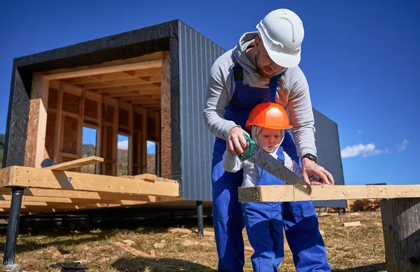 Father with toddler son building wooden frame house in the Scandinavian style barnhouse. Male builders using hand saw to cut boards on construction site on sunny day. Carpentry and family concept.