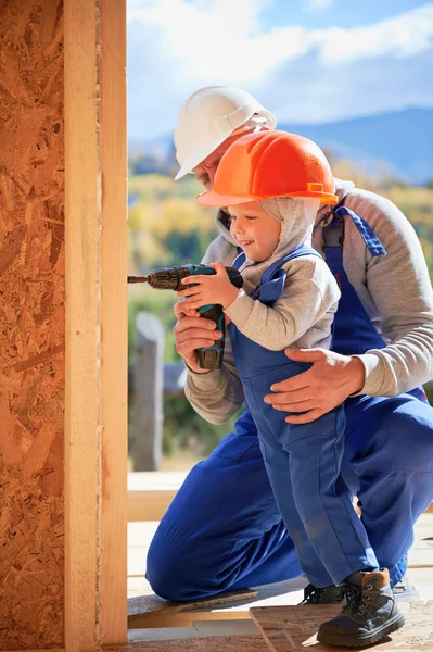Father with toddler son building wooden frame house. Male builder and kid using screwdriver on construction site, wearing helmet and blue overalls on sunny day. Carpentry and family concept.