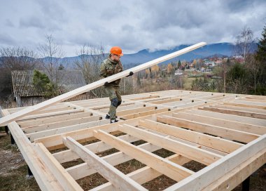 Man worker building wooden frame house on pile foundation. Carpenter carrying wooden board. Carpentry concept.