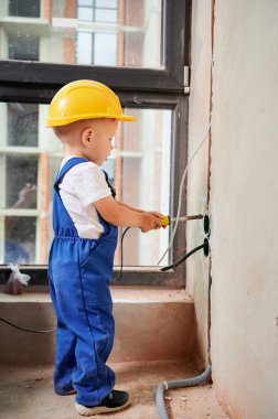 Kid welding electrical wire with electric soldering iron. Child electrician in work overalls and safety helmet installing electrical cables and sockets in apartment under renovation.