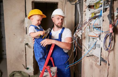 Child electrician standing in ladder and holding pliers tool while installing switchboard with father at home. Man and kid mounting electrical wiring in apartment under renovation.