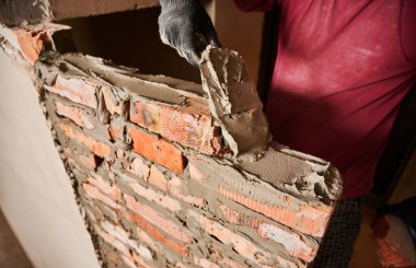 Close up of man hand in work glove laying brickwork in building under construction. Male worker applying cement mortar on bricks with trowel tool. Masonry construction concept.