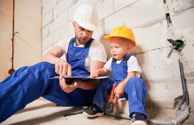 Male worker sitting by brick wall and pointing at clipboard while little boy holding tool. Man and kid wearing safety helmets and work overalls while working with documents at construction site.