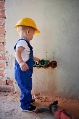 Little boy electrician installing cable canals and sockets in wall. Child in safety and work overalls construction helmet mounting electrical wiring in apartment under renovation.