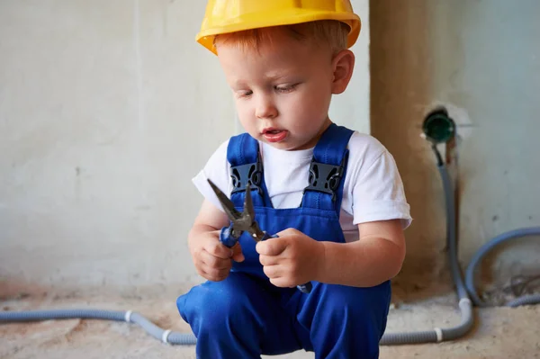 Adorable baby boy looking at instrument in building under construction. Cute child wearing work overalls and safety helmet while playing with tool at construction site.