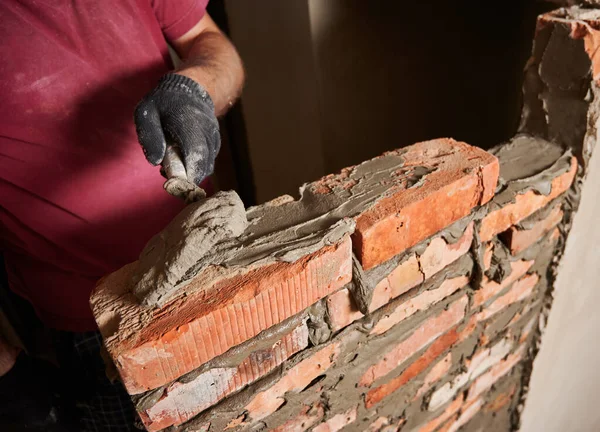 Close up of male hand in work glove laying brickwork in building under construction. Man bricklayer builder applying cement mortar on bricks with trowel tool. Masonry construction concept.