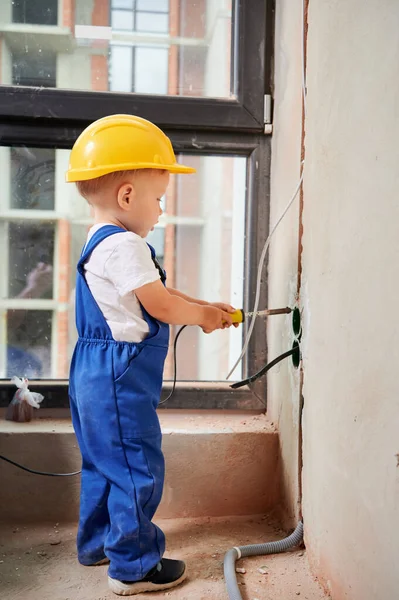 Kid welding electrical wire with electric soldering iron. Child electrician in work overalls and safety helmet installing electrical cables and sockets in apartment under renovation.