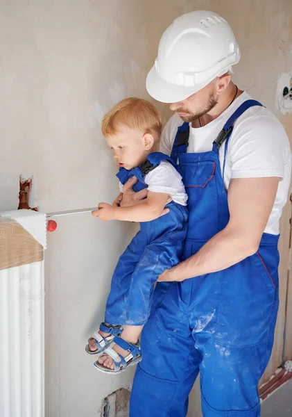 Adorable child using construction tool while installing heating radiator with father at home. Male plumber in work overalls and safety helmet holding baby boy. Home renovation and parenting concept.