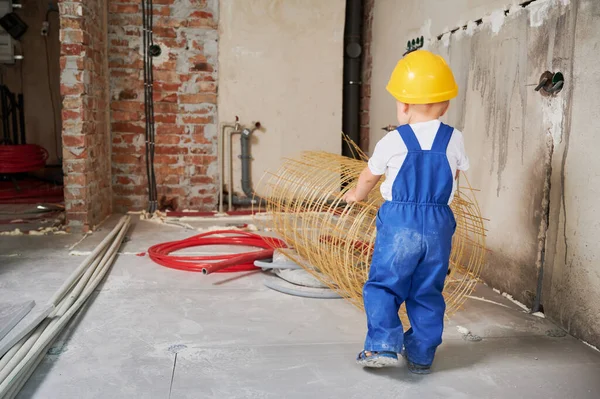 Back view of baby boy in safety helmet using mesh fiberglass rebar for home renovation. Child builder in work overalls standing by construction materials in apartment room with pipes on the floor.
