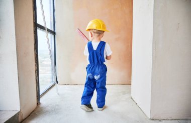Child checking wall surface with spirit level in apartment. Back view of kid in work overalls and safety construction helmet using level instrument while working on home renovation.