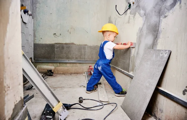 Boy construction worker checking wall surface with spirit level tool in apartment. Kid in work overalls using level instrument while preparing wall for repair works at home during renovation.