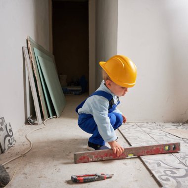 Child construction worker holding spirit level instrument. Kid in safety helmet and work overalls playing in apartment under renovation.