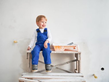 Cute kid construction worker sitting on wooden table against white wall in apartment under renovation. Cheerful little boy wearing safety helmet and work overalls while playing at home.