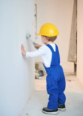 Full length of child construction worker painting wall in apartment under renovation. Kid in safety helmet and work overalls using paint roller while working on home renovation.
