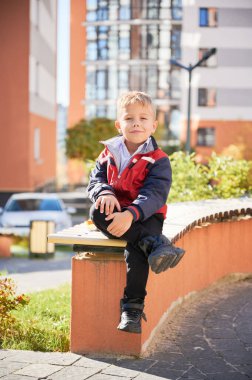 Cute boy in casual wear smiling, while sitting on bench with cross legs outdoors. Portrait view of joyful kid putting arms on knees at sunny day. Modern residential buildings on background.