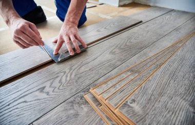 Man preparing laminate plank for floor installation in apartment under renovation. Close up of male worker using metal construction ruler and pen while drawing line on laminate flooring board.