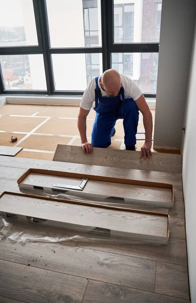 Male construction worker laying laminate wood plank on floor underlayment. Man in work overalls installing laminate timber flooring in room with large window. Hardwood floor renovation concept.