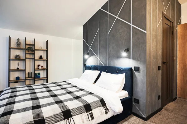 Bedroom with big bed and decorated wooden shelves indoors. Side view of wide bed, with checkered plaid against gray wall with geometric pattern in bedroom. Concept of interior.