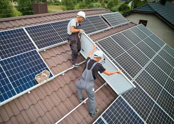 Men workers mounting photovoltaic solar moduls on roof of house. Engineers in helmet installing solar panel system outdoors. Concept of alternative and renewable energy. Aerial view.