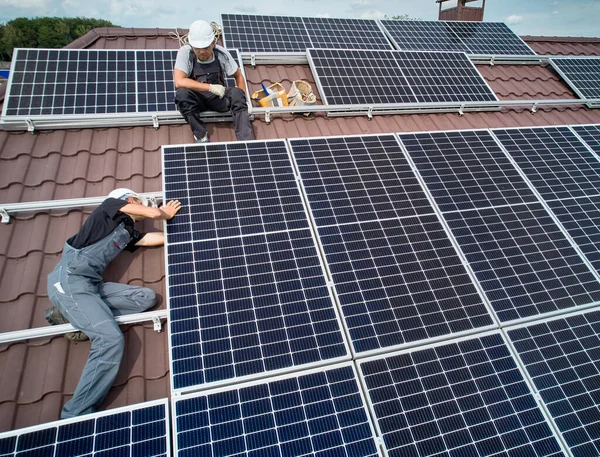 Men technicians mounting photovoltaic solar moduls on roof of house. Workers in helmets installing solar panel system outdoors. Concept of alternative and renewable energy. Aerial view.