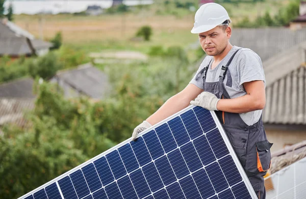 Portrait of man worker mounting photovoltaic solar moduls on roof of house. Electrician in helmet installing solar panel system outdoors. Concept of alternative and renewable energy.