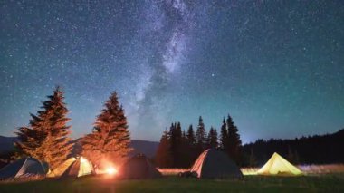Timelapse of the starry sky with the Milky Way. Night camping with five tourists illuminated tents. Hiker having a rest near campfire and forest, cooking dinner on fire. Concept of tourism and nature.