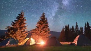 Night camping with five tourists illuminated tents. Hiker having a rest near campfire and forest, cooking dinner on fire. Timelapse of the starry sky with the Milky Way. Concept of tourism and nature.