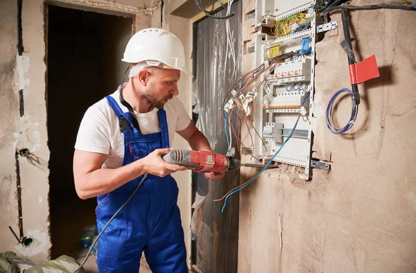 Male builder installing electrical switch panel in apartment. Man in work overalls and safety helmet drilling wall with power tool electric drill while standing by the wall with switchboard.