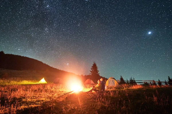 Night camping in mountains under starry sky. Two people, couple sitting on grass near burning campfire, enjoying beautiful sky full of stars. Concept of tourism and healthy living.