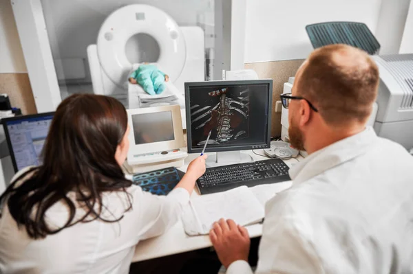 Medical computed tomography or MRI scanner. Back view of two doctors sitting, working at computer, examining MRI results. Specialists woman pointing by pen, patient lying. Concept of medicine.