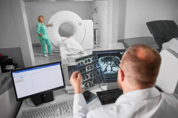 Medical computed tomography or MRI scanner. Back view of male doctor sitting at computer, nurse making MRI. Focus on nurse. Specialists working in hospital, studying results, examining.