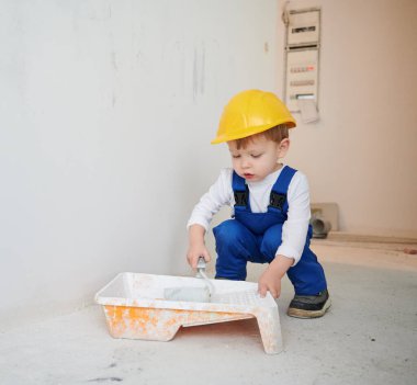 Adorable child construction worker crouching down and preparing paint roller for wall painting. Kid wearing safety helmet and work overalls while working on home renovation.