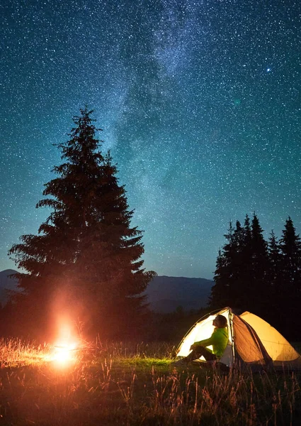 Night camping in mountains under starry sky and Milky way. Female tourist sitting in tent entrance in campsite, admiring landscape and burning campfire. Concept of traveling and hiking.