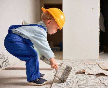Side view of little boy construction worker using brush while playing at home. Kid wearing safety helmet and work overalls while preparing floor for renovation works in apartment.