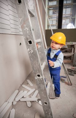 Little boy construction worker with ladder while working on home renovation. Kid in safety helmet and work overalls playing in apartment under renovation.