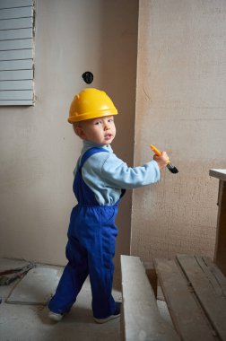 Boy construction worker painting wall with brush in apartment under renovation. Child wearing safety helmet and work overalls while playing at home.