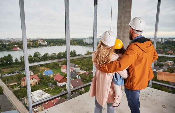 Back view of parents with daughter standing inside apartment building under construction. Man and woman with child wearing building helmets while enjoying city view from future home.