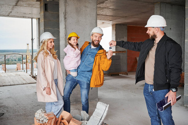 Male builder handing keys from new apartment to family with child at construction site. Happy parents with little daughter receiving keys to new home from construction worker.