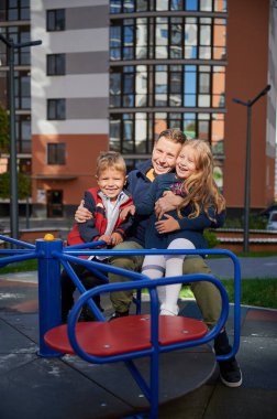 Mutlu baba, şehrin yüksek binalarının modern avlusunda iki çocuğa sarılıyor. Ebeveynlik kavramı.