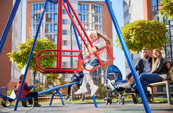 stock image Little daughter playing on swing, while mum and dad sitting on bench at playground. Side view of adorable girl on swing smiling, while parents looking after child behind at daytime. Concept of joy.