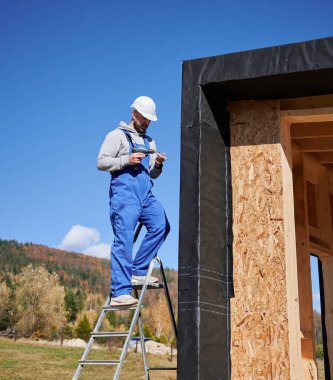 Male builder installing black corrugated iron sheet used as facade of future cottage, blue sky on background. Man worker building wooden frame house. Carpentry and construction concept.