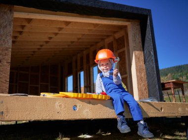 Boy toddler playing as builder on construction site. Child carpenter in orange helmet and blue overalls learning to build wooden frame house outdoor on sunny day. Carpentry and workshop concept.