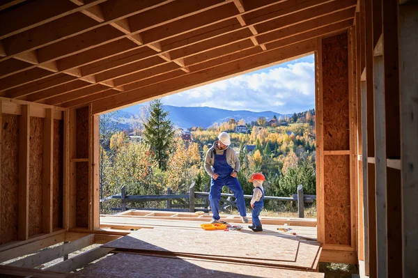 Father with toddler son building wooden frame house in the Scandinavian style barnhouse. Male builder having fun with kid on construction site on sunny day. Carpentry and family concept.