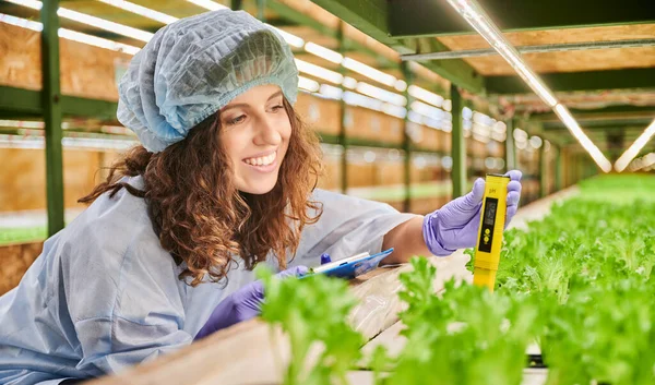 Joyful female gardener in disposable cap holding clipboard and smiling while measuring soil pH value with digital electronic device. Woman scientist using pH meter in greenhouse
