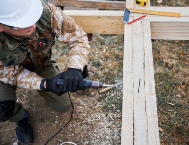 Yığınla temele ev inşa eden bir adam. Erkek işçilerin gelecekteki evin ahşap çerçevesinde elektrikli matkapla delik açtıkları yeri kapatın. Beyaz kask takan bir mimar. Marangozluk kavramı.