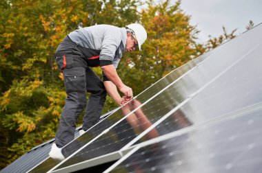 Man electrician mounting photovoltaic solar panels on roof of house. Engineer in helmet installing solar module system with help of hex key. Concept of alternative, renewable energy. clipart