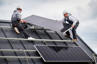 Teknisyenler evin çatısına güneş paneli kuruyor. Dışarıda fotovoltaik güneş modülü taşıyan miğferli işçiler. Alternatif ve yenilenebilir enerji kavramı.